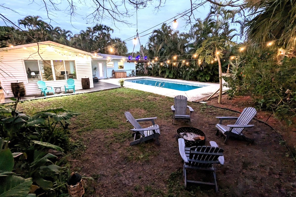 Backyard of a vacation rental at dusk with a pool, Adirondack chairs around a fire pit, string lights, and tropical palms