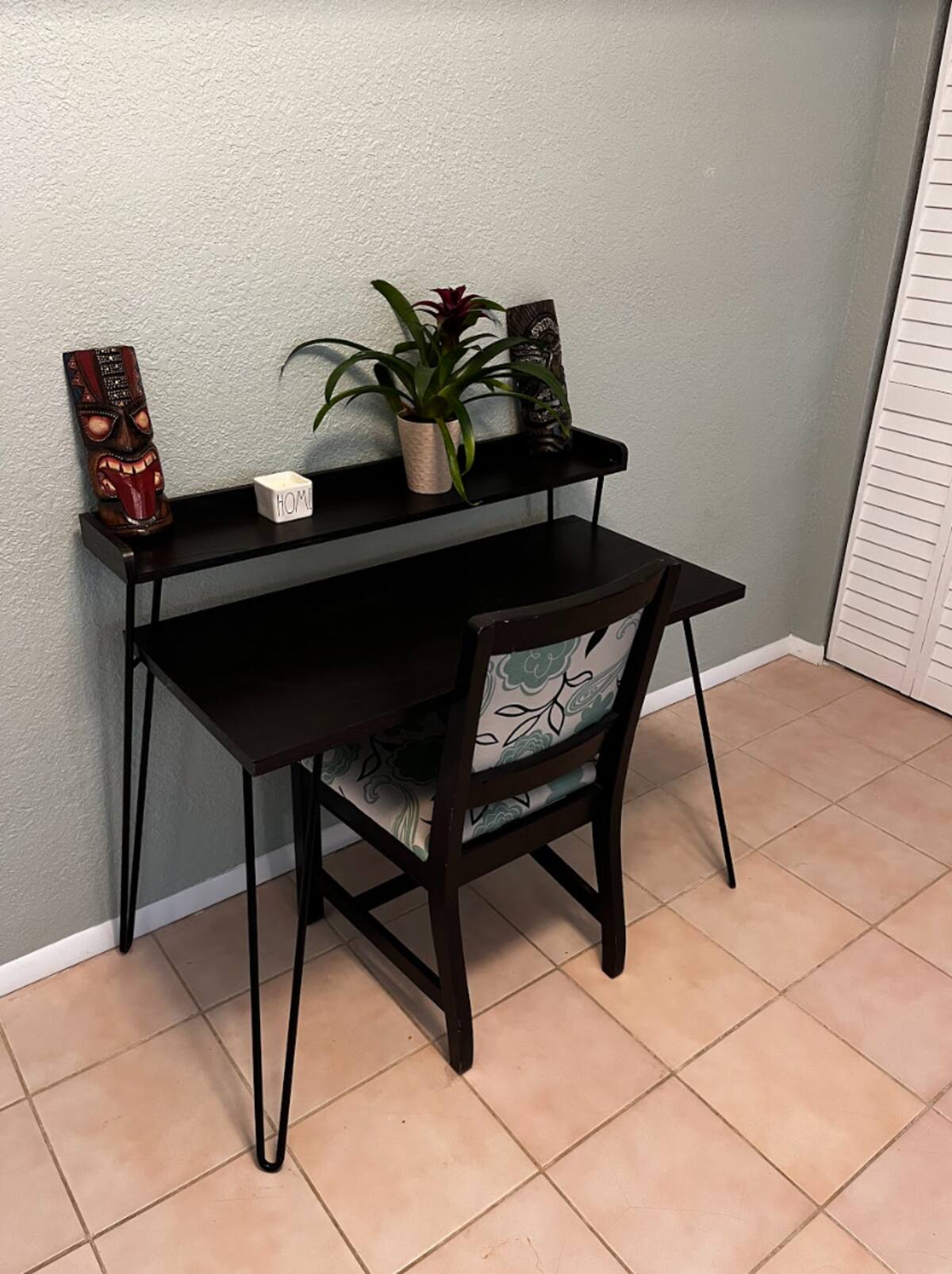 Small dark wood desk and floral-upholstered chair with tiki decor and a plant against a gray wall in a tiled room