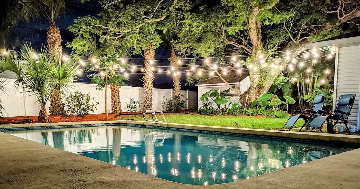 Private backyard pool at night, lit by string lights strung between mature palms and shade trees
