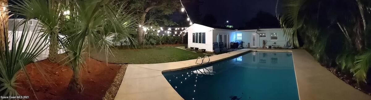 Backyard pool at night with string lights and palm trees behind a single-story house