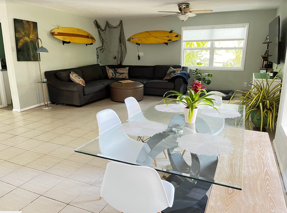 Bright beach-themed living and dining area with sectional sofa, surfboard wall decor, and glass dining table