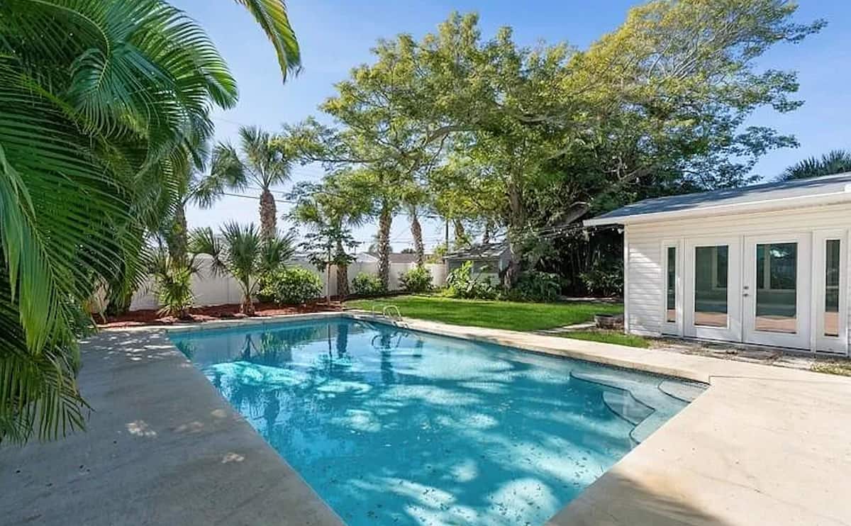 Backyard swimming pool surrounded by palm trees with a white poolside house and French doors