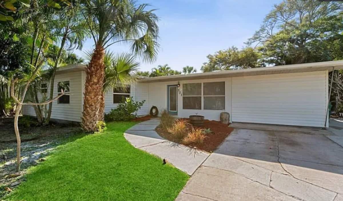 Single-story white vacation rental home with palm trees, green lawn, and landscaped entrance under a blue sky